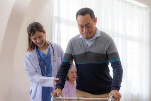 asiatisch jung Krankenschwester Unterstützung Senior älter Mann gehen mit Gehhilfe. schön Krankenschwester Hilfe und nehmen Pflege. Alten reifen Opa geduldig tun physisch Therapie mit ein Gruppe von Senior freunde im Krankenhaus. foto