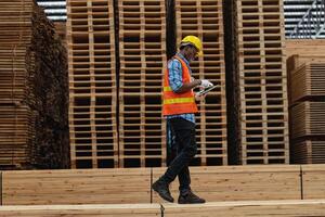 afrikanisch Arbeitskräfte Mann Ingenieurwesen Gehen und inspizieren mit Arbeiten Suite Kleid und Hand Handschuh im Bauholz Holz Lagerhaus. Konzept von Clever Industrie Arbeiter Betriebs. Holz Fabriken produzieren Holz Gaumen. foto