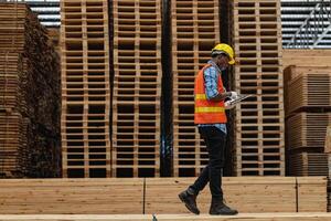 afrikanisch Arbeitskräfte Mann Ingenieurwesen Gehen und inspizieren mit Arbeiten Suite Kleid und Hand Handschuh im Bauholz Holz Lagerhaus. Konzept von Clever Industrie Arbeiter Betriebs. Holz Fabriken produzieren Holz Gaumen. foto