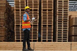 afrikanisch Arbeitskräfte Mann Ingenieurwesen Gehen und inspizieren mit Arbeiten Suite Kleid und Hand Handschuh im Bauholz Holz Lagerhaus. Konzept von Clever Industrie Arbeiter Betriebs. Holz Fabriken produzieren Holz Gaumen. foto