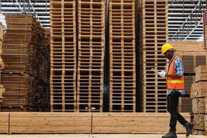 afrikanisch Arbeitskräfte Mann Ingenieurwesen Gehen und inspizieren mit Arbeiten Suite Kleid und Hand Handschuh im Bauholz Holz Lagerhaus. Konzept von Clever Industrie Arbeiter Betriebs. Holz Fabriken produzieren Holz Gaumen. foto