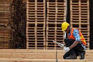 afrikanisch Arbeitskräfte Mann Ingenieurwesen Gehen und inspizieren mit Arbeiten Suite Kleid und Hand Handschuh im Bauholz Holz Lagerhaus. Konzept von Clever Industrie Arbeiter Betriebs. Holz Fabriken produzieren Holz Gaumen. foto