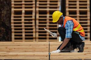 afrikanisch Arbeitskräfte Mann Ingenieurwesen Gehen und inspizieren mit Arbeiten Suite Kleid und Hand Handschuh im Bauholz Holz Lagerhaus. Konzept von Clever Industrie Arbeiter Betriebs. Holz Fabriken produzieren Holz Gaumen. foto