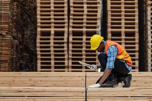 afrikanisch Arbeitskräfte Mann Ingenieurwesen Gehen und inspizieren mit Arbeiten Suite Kleid und Hand Handschuh im Bauholz Holz Lagerhaus. Konzept von Clever Industrie Arbeiter Betriebs. Holz Fabriken produzieren Holz Gaumen. foto