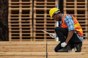 afrikanisch Arbeitskräfte Mann Ingenieurwesen Gehen und inspizieren mit Arbeiten Suite Kleid und Hand Handschuh im Bauholz Holz Lagerhaus. Konzept von Clever Industrie Arbeiter Betriebs. Holz Fabriken produzieren Holz Gaumen. foto