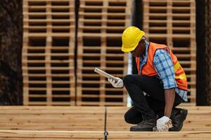 afrikanisch Arbeitskräfte Mann Ingenieurwesen Gehen und inspizieren mit Arbeiten Suite Kleid und Hand Handschuh im Bauholz Holz Lagerhaus. Konzept von Clever Industrie Arbeiter Betriebs. Holz Fabriken produzieren Holz Gaumen. foto