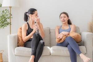 Gruppe von glücklich sportlich Frauen tragen Körper stilvoll Sportbekleidung und reden zusammen. warten zum Yoga Klasse.. gesund Lebensstil und Wohlbefinden. Paar Frauen sind entspannend und Sitzung beim Sofa im Leben Zimmer. foto