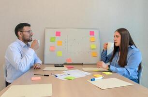 ein Mann und Frau Sitzung beim ein Tabelle mit ein Weiß Tafel und klebrig Anmerkungen foto