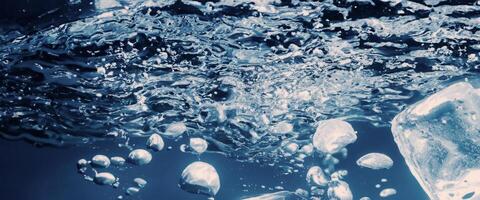 Wasser planschen und Eis Würfel. Eis planschen in ein Glas von Wasser. unter Wasser Gießen Eis Würfel fallen in klar Bewässerung Hintergrund. erfrischend Ausruhen Trinken. Eis im ein Glas mit Blau Hintergrund foto