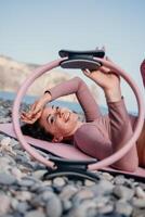 Frau Meer Pilates. sportlich glücklich Mitte alt Frau üben Fitness auf Strand in der Nähe von Meer, lächelnd aktiv weiblich Ausbildung mit Ring auf Yoga Matte außen, genießen gesund Lebensstil, Harmonie und Meditation foto