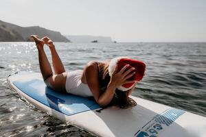 Frau Meer sup. schließen oben Porträt von glücklich jung kaukasisch Frau mit lange Haar im Santa Hut suchen beim Kamera und lächelnd. süß Frau Porträt im ein Weiß Bikini posieren auf sup Tafel im das Meer foto