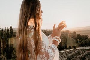 glücklich Frau im Weiß Boho Kleid auf Sonnenuntergang im Berge. romantisch Frau mit lange Haar Stehen mit ihr zurück auf das Sonnenuntergang im Natur im Sommer- mit öffnen Hände. Silhouette. Natur. Sonnenuntergang. foto