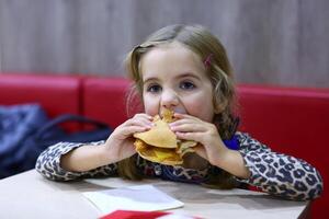 Porträt von ein kaukasisch vier Jahr alt Mädchen Essen Burger im ein Cafe foto