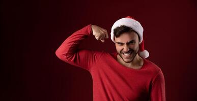 jung gut aussehend kaukasisch Kerl im ein rot Sweatshirt und Santa Hüte steht auf rot Hintergrund im Studio und zeigen Bizeps Leistung foto