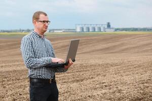 modern Farmer Überprüfung seine Feld Pflanze und Arbeiten auf Laptop Computer gegen Mais Trockner Silos im Konzept von industriell und Landwirtschaft foto