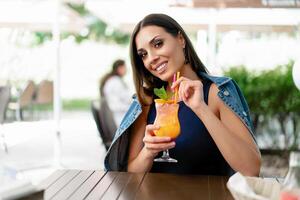ein schön jung Mädchen von europäisch Aussehen ist Trinken ein exotisch Sommer- Cocktail während Sitzung beim ein Tabelle von ein Straße Sommer- Cafe foto