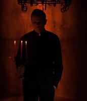 jung katholisch beten Priester. Porträt von Priester Nächster zu das Kerzen betet mit seine Hände gefaltet in der Nähe von das Gesicht. rot Mauer auf das Hintergrund. Innen- gut aussehend Porträt von Religion Mann foto