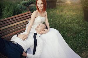 Bräutigam Lügen auf das schön Mädchen im ein Hochzeit Kleid im das Öffentlichkeit Park auf das Bank foto