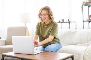 Frau Brille mit Laptop Computer während Sitzung Sofa mit groß Fenster auf Hintergrund beim Zuhause Innere foto