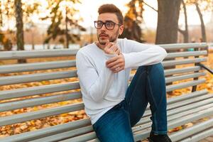 jung gut aussehend Hipster Kerl ist Sitzung im Herbst Park auf ein Bank. gelangweilt allein. ein Schüler Spaziergänge im das Park. foto