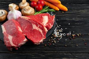 drei Stücke von saftig roh Rindfleisch mit Gemüse auf ein Stein Schneiden Tafel auf ein schwarz hölzern Tabelle Hintergrund. foto