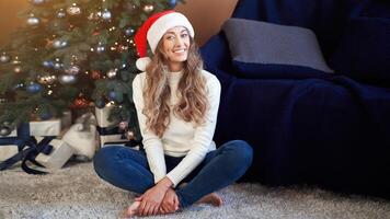 Weihnachten. Frau gekleidet Weiß Sweatshirt und Jeans Sitzung auf das Fußboden in der Nähe von Weihnachten Baum mit Geschenk Box foto