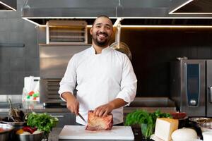 gut aussehend jung afrikanisch Koch Stehen im Fachmann Küche im Restaurant vorbereiten ein Mahlzeit von Fleisch und Käse Gemüse. foto