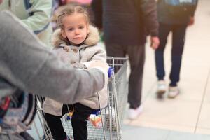 wenig Mädchen Sitzung im ein Supermarkt Wagen und suchen im zu das Kamera foto