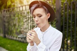 schön informell Mädchen mit lila kurz Haar und im ein modisch Hut raucht ein Zigarette foto