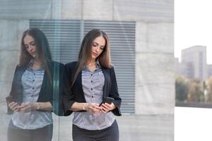 Geschäft Frau 35 Jahre gekleidet Streifen Hemd und schwarz Jacke mit lange Haar Stehen in der Nähe von Büro Gebäude draussen verwenden Smartphone foto