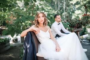 ein schön Braut im ein Weiß Hochzeit Kleid und Kranz sitzt auf ein Stuhl Nächster zu das Bräutigam, ruhen und vorbereiten zum ein glücklich Familie Leben foto