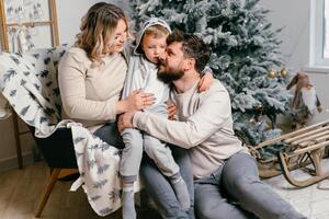 Weihnachten Familie Glück Porträt von Papa, schwanger Mama und wenig Sohn Sitzung Sessel beim Zuhause in der Nähe von Weihnachten Baum Umarmung Lächeln foto