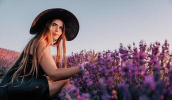 Frau Lavendel Feld. glücklich sorglos Frau im schwarz Kleid und Hut mit groß Rand Gehen im ein Lavendel Feld während Sonnenuntergang. perfekt zum inspirierend und warm Konzepte im Reise und Fernweh. foto