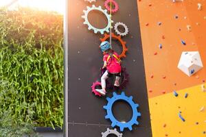 Kind Klettern auf Mauer im Amüsement Center. Klettern Ausbildung zum Kinder. wenig Mädchen im gekleidet Klettern Ausrüstung steigen hoch. extrem aktiv Freizeit zum Kinder. foto