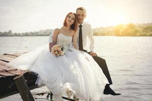 Hochzeit Paar Sitzung auf Brücke in der Nähe von See auf Sonnenuntergang beim Hochzeit Tag. Braut und Bräutigam im Liebe foto