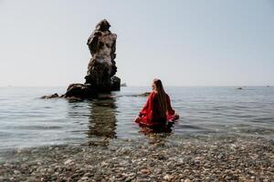 Frau Reise Meer. glücklich Tourist im rot Kleid genießen nehmen Bild draußen zum Erinnerungen. Frau Reisender posieren auf das Felsen beim Meer Bucht umgeben durch vulkanisch Berge, Teilen Reise Abenteuer Reise foto