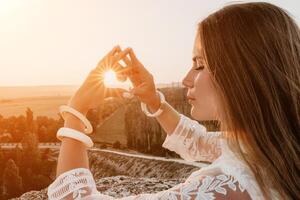 glücklich Frau im Weiß Boho Kleid Herstellung Herz Zeichen mit Hände auf Sonnenuntergang im Berge. romantisch Frau mit lange Haar Stehen mit ihr zurück auf das Sonnenuntergang im Natur im Sommer- mit öffnen Hände. foto