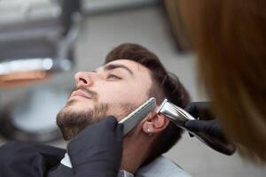 jung schön kaukasisch Frau Friseur schneidet Bart gut aussehend Mann beim modern Friseur foto