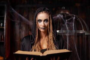 Halloween Konzept. Hexe gekleidet schwarz Kapuze mit Dreadlocks Stehen dunkel Verlies Zimmer verwenden Magie Buch zum beschwören Magie buchstabieren foto