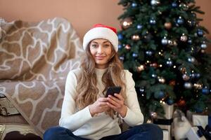 Weihnachten. Frau gekleidet Weiß Sweatshirt Santa Hut auf das Fußboden in der Nähe von Weihnachten Baum mit Smartpone und Tasse von Kaffee foto