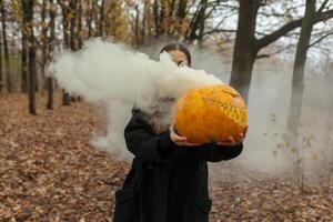 ein schön Mädchen mit lange Haar im ein schwarz Mantel ist halten ein Orange Kürbis von welche Rauch ist Kommen. glücklich Halloween. dunkel Herbst Wald. Schrecken foto