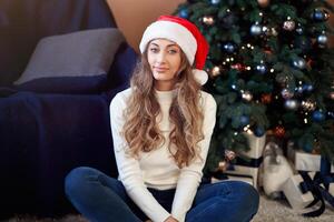 Weihnachten. Frau gekleidet Weiß Sweatshirt und Jeans Sitzung auf das Fußboden in der Nähe von Weihnachten Baum mit Geschenk Box foto