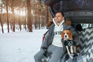 ein jung Familie von Reisende ein Kerl sind Sitzung im das Kofferraum von ihr Auto im ein Umarmung mit ihr Freund Hund Beagle. Winter Wald auf das Hintergrund. Reisen mit ein Hund durch Auto foto