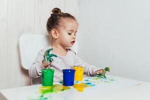 schön wenig Mädchen zeichnet mit Finger Farben auf ein Weiß Blatt von Papier. kreativ Kind Entwicklung im Kindergarten oder kostenlos Zeit beim Zuhause foto