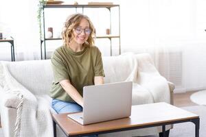 Frau Brille mit Laptop Computer während Sitzung Sofa mit groß Fenster auf Hintergrund beim Zuhause Innere foto