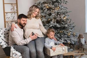 Weihnachten Familie Glück Porträt von Papa, schwanger Mama und wenig Sohn Sitzung Sessel beim Zuhause in der Nähe von Weihnachten Baum Umarmung Lächeln foto