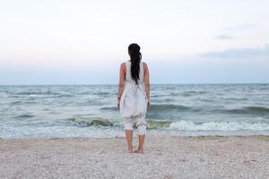 zurück Aussicht von schön Frau im Weiß Kleid mit Dreadlock auf das Kopf genießen das idyllisch Szene auf das Strand. foto