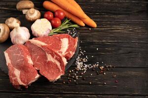 drei Stücke von saftig roh Rindfleisch mit Gemüse auf ein Stein Schneiden Tafel auf ein schwarz hölzern Tabelle Hintergrund. foto