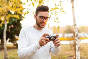 jung gut aussehend Hipster Kerl Spaziergänge im ein schön Herbst Park auf das Hintergrund von Gelb Blätter im warm sonnig Wetter und nimmt Bilder auf ein Schwein Film Kamera. Herbst Freizeit Zeit. kreativ Jugend foto