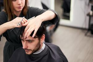 gut aussehend Blau Augen Mann Sitzung im Barbier Geschäft. Friseur Friseur Frau Schneiden seine Haar. weiblich Barbier. foto